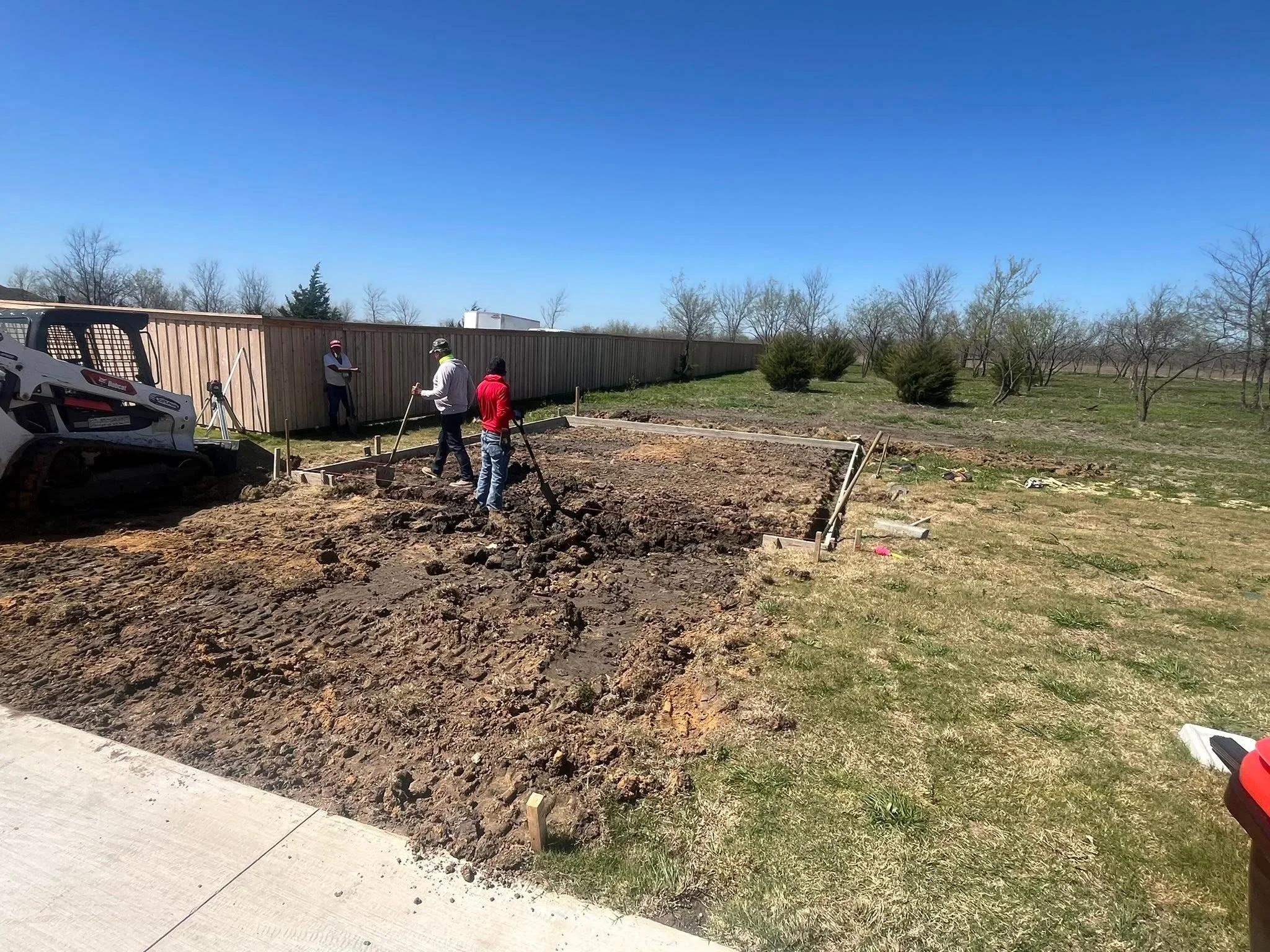 Excavated fence line with wooden stakes marking post locations on prepared soil