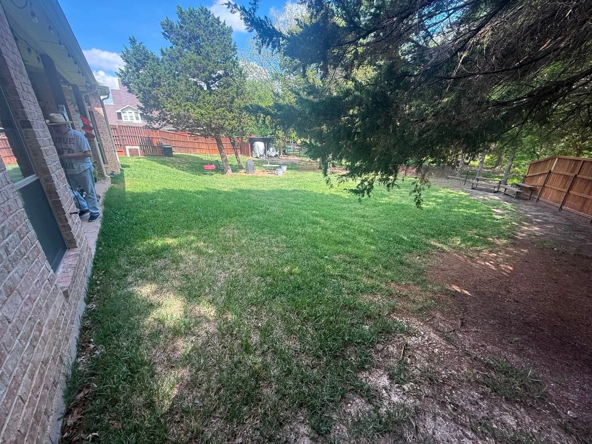 Backyard with green lawn, wooden fencing, mature trees, and brick home exterior
