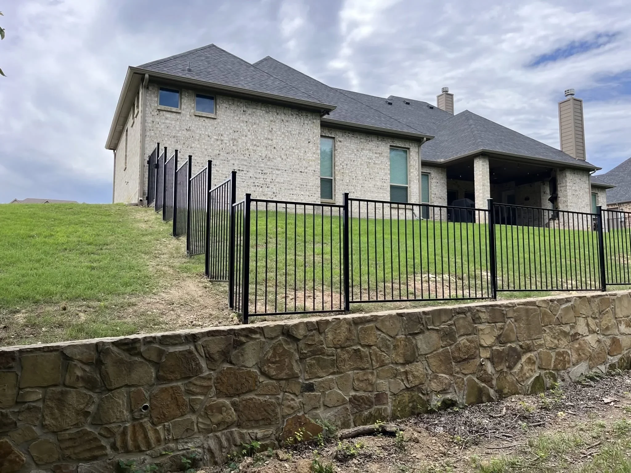 Black aluminum picket fence with vertical spindles installed on sloped terrain with stone retaining wall