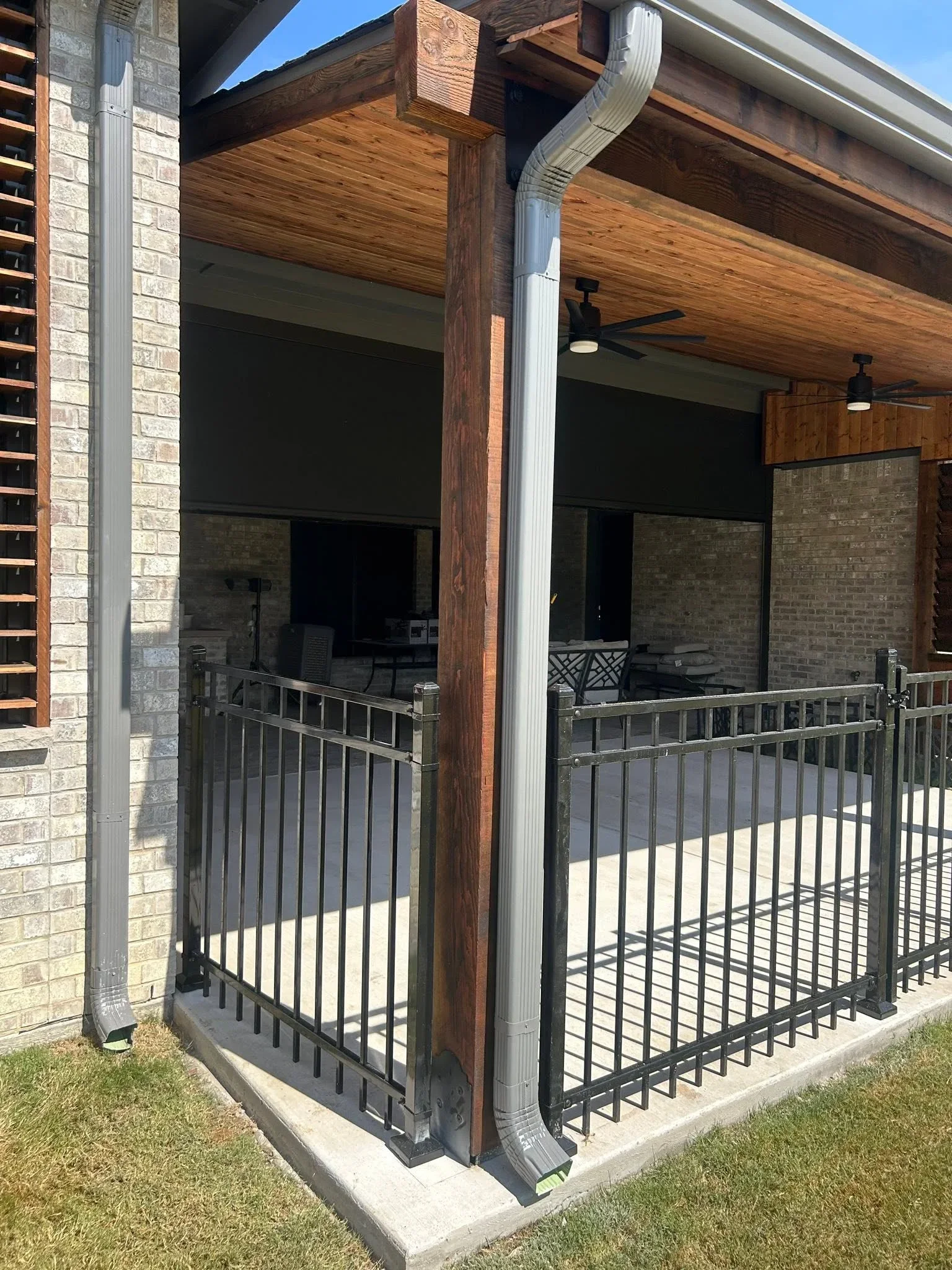 Black metal railing with vertical pickets, timber pergola beams, brick columns, and gutter system