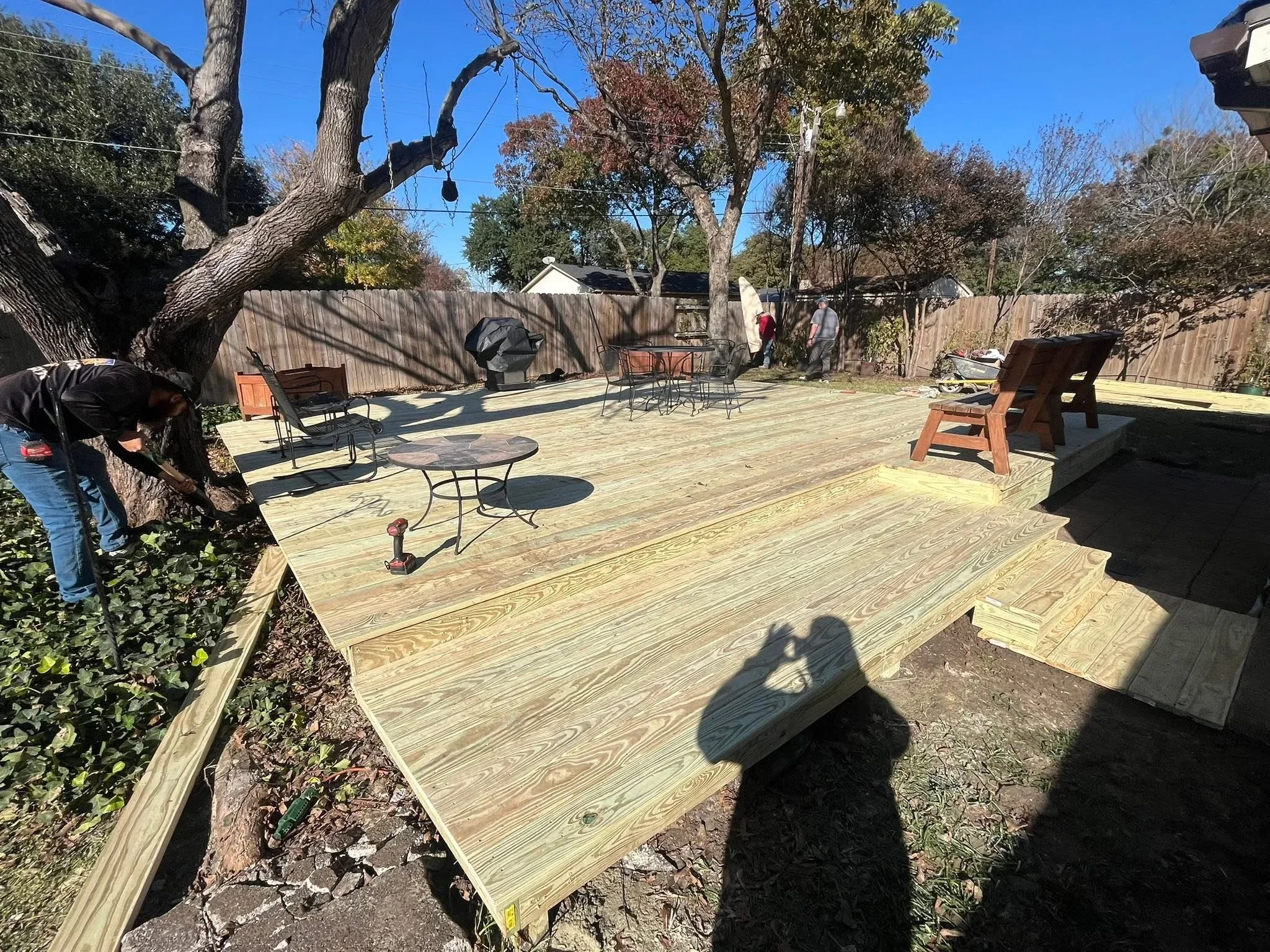 Composite deck with multiple levels built around mature trees, metal patio furniture, and wooden fence backdrop