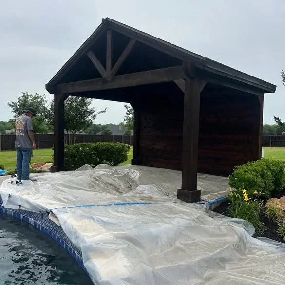 Dark stained wood pavilion with exposed beam trusses over concrete patio near pool
