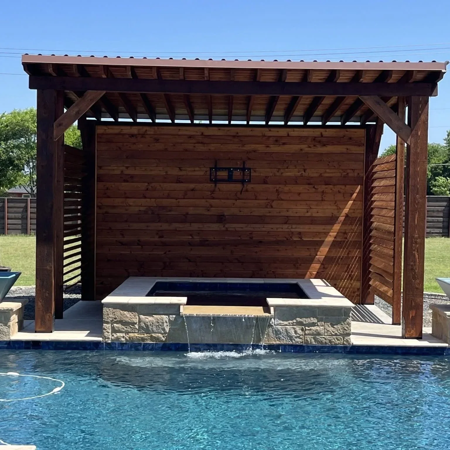 Dark wood pergola with stone fire pit and waterfall feature beside swimming pool