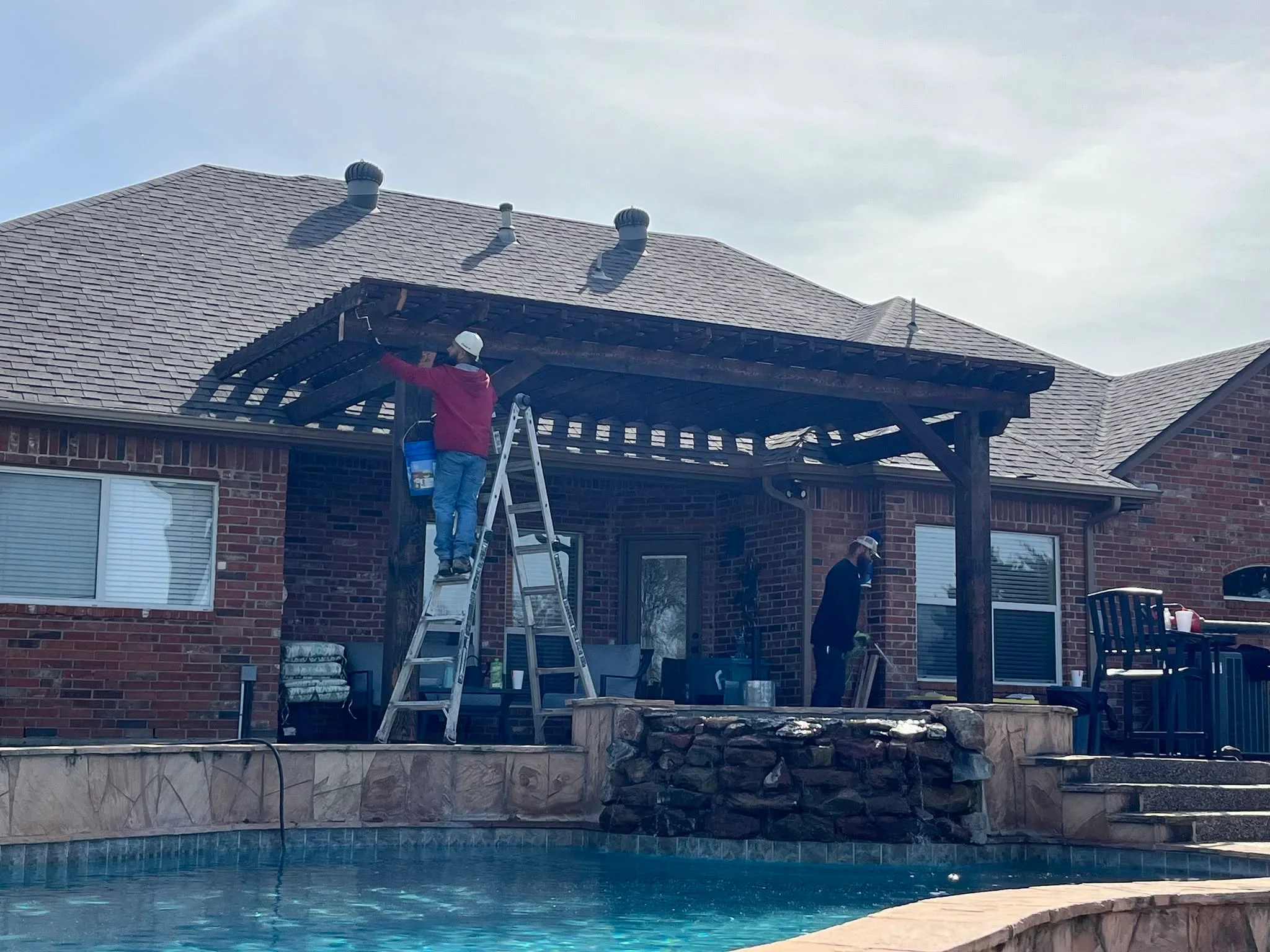 Dark wood pergola with slatted roof installed over pool patio adjacent to brick home