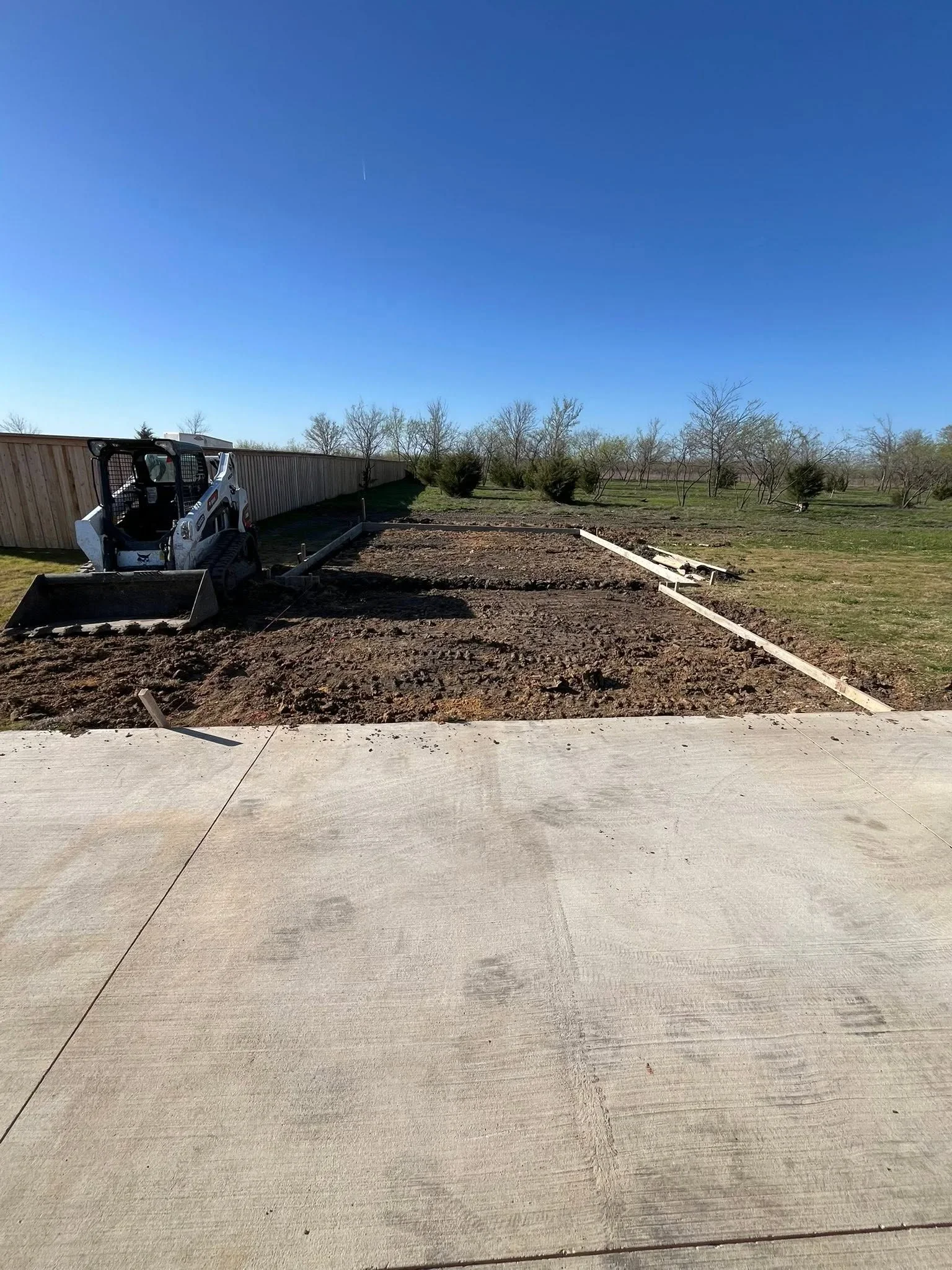 Concrete driveway with control joints, skid steer loader, graded soil, and wooden perimeter framing