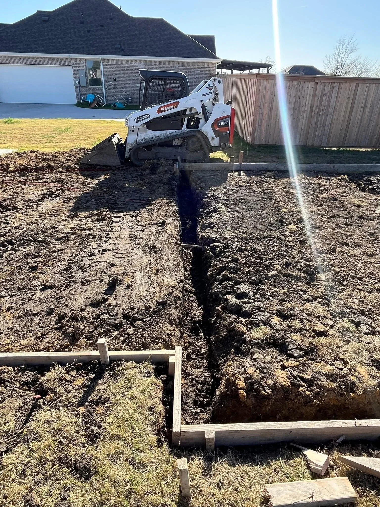 Bobcat excavator grading soil trench for fence post installation with wooden frame borders