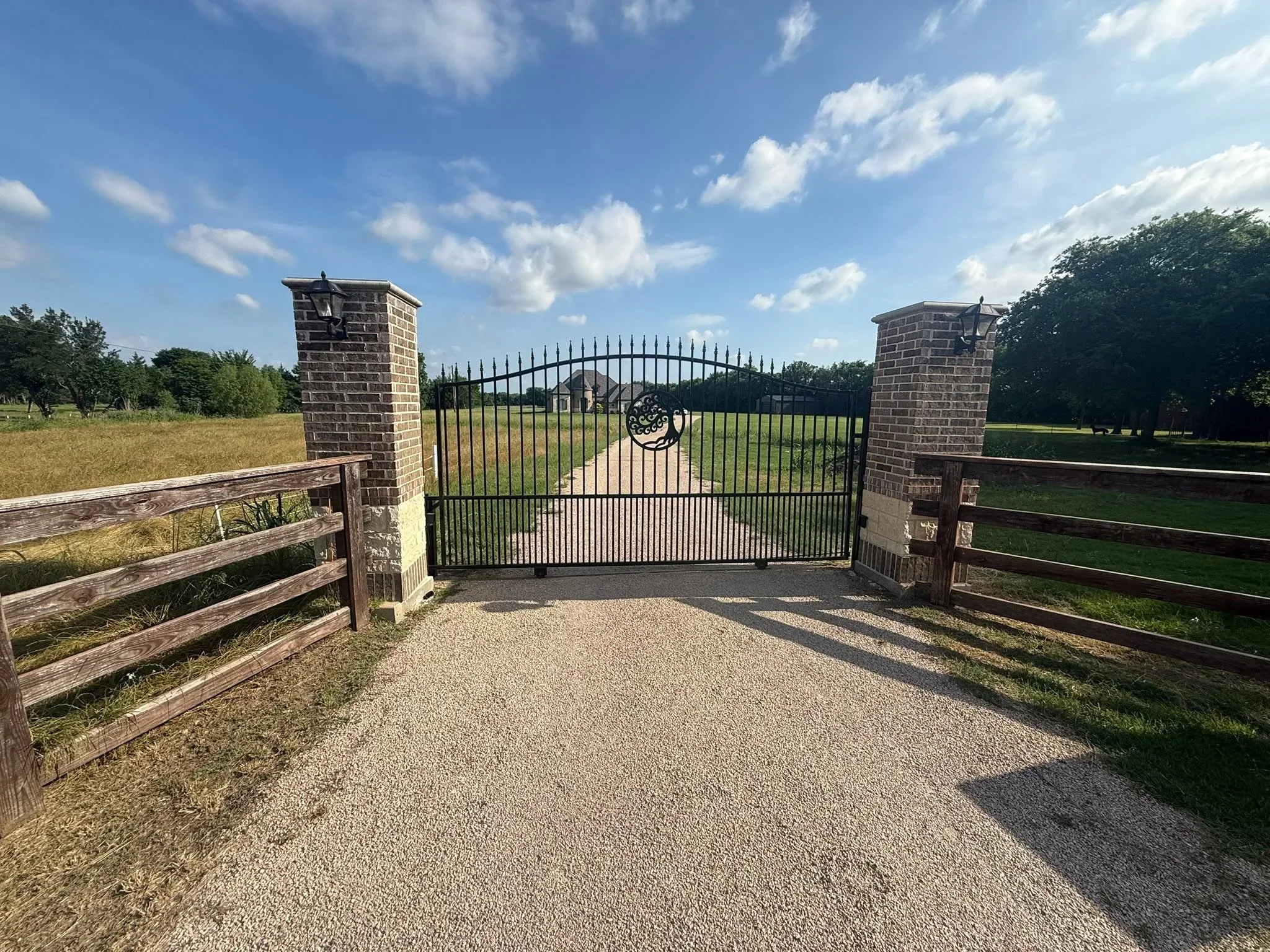 Black metal arched driveway gate between brick pillars with wooden ranch fencing on gravel drive