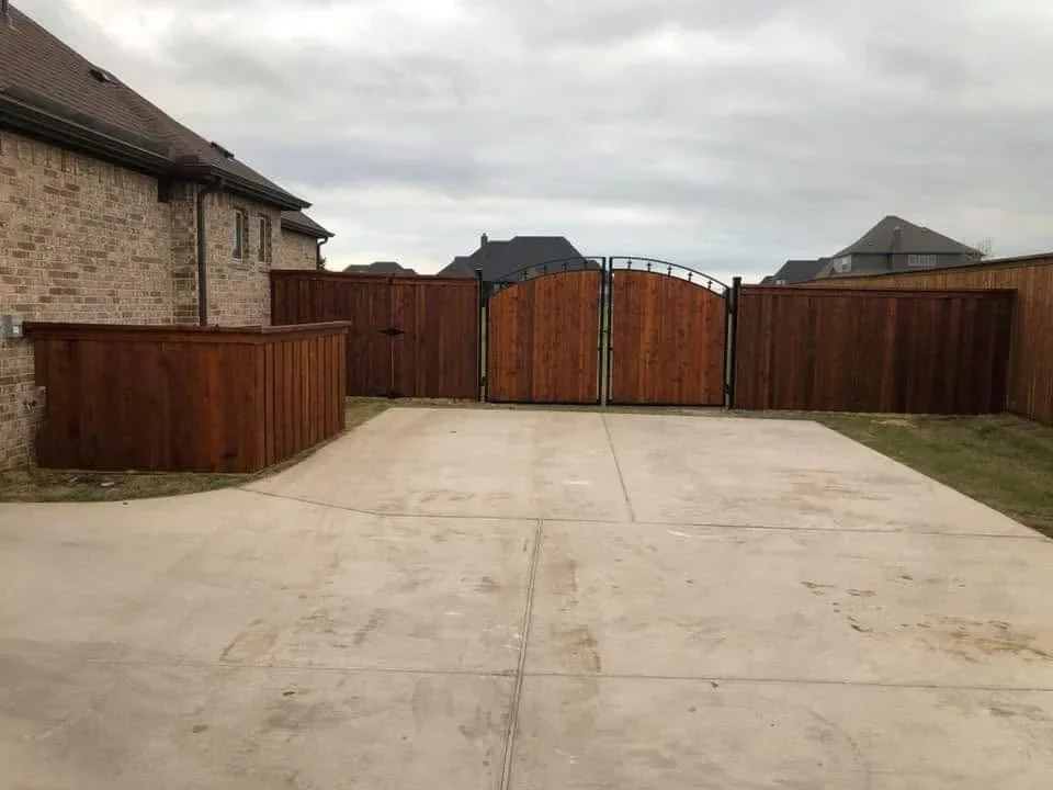 Stained wood privacy fence with arched gate and metal frame accents along concrete driveway