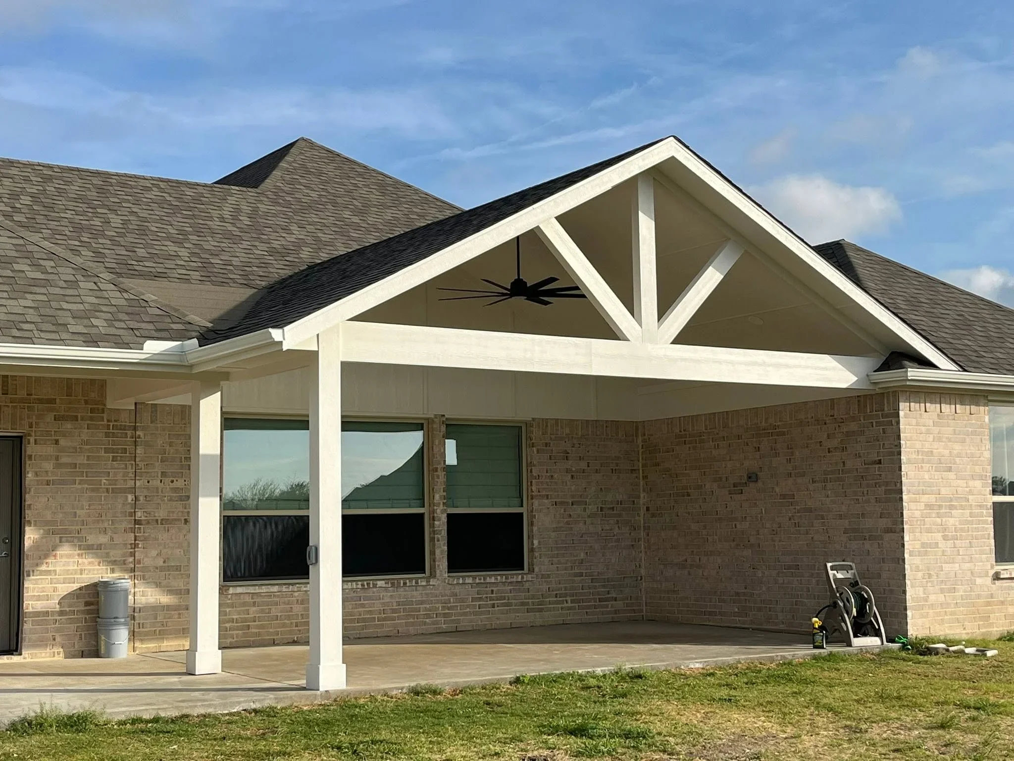 White covered patio with gable roof, decorative beams, and concrete pad attached to brick home