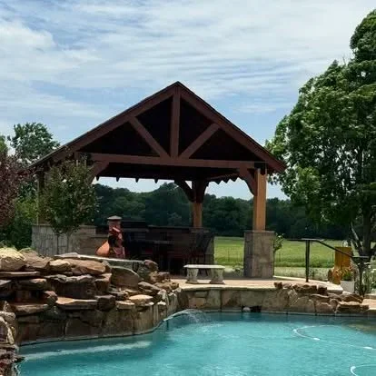 Timber pavilion with exposed beams and gable roof overlooking pool with stone fireplace