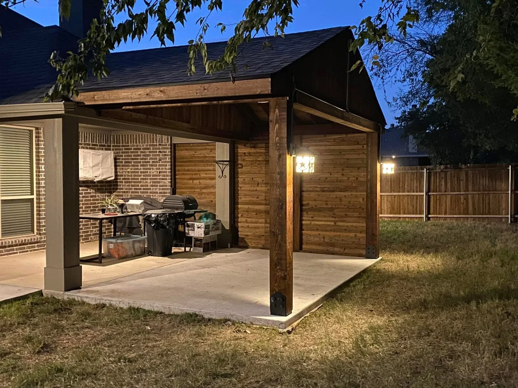 Cedar pergola with vertical wood panels, concrete patio, and grill area at dusk with landscape lighting