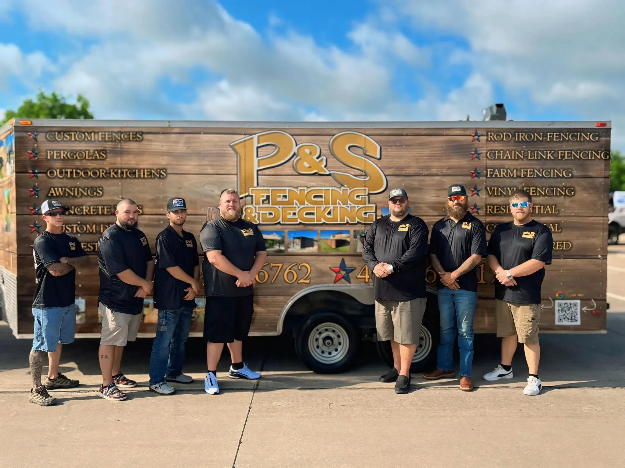 Team of seven workers in black company shirts standing in front of wood-wrapped service trailer with P&S Fencing and Decking branding