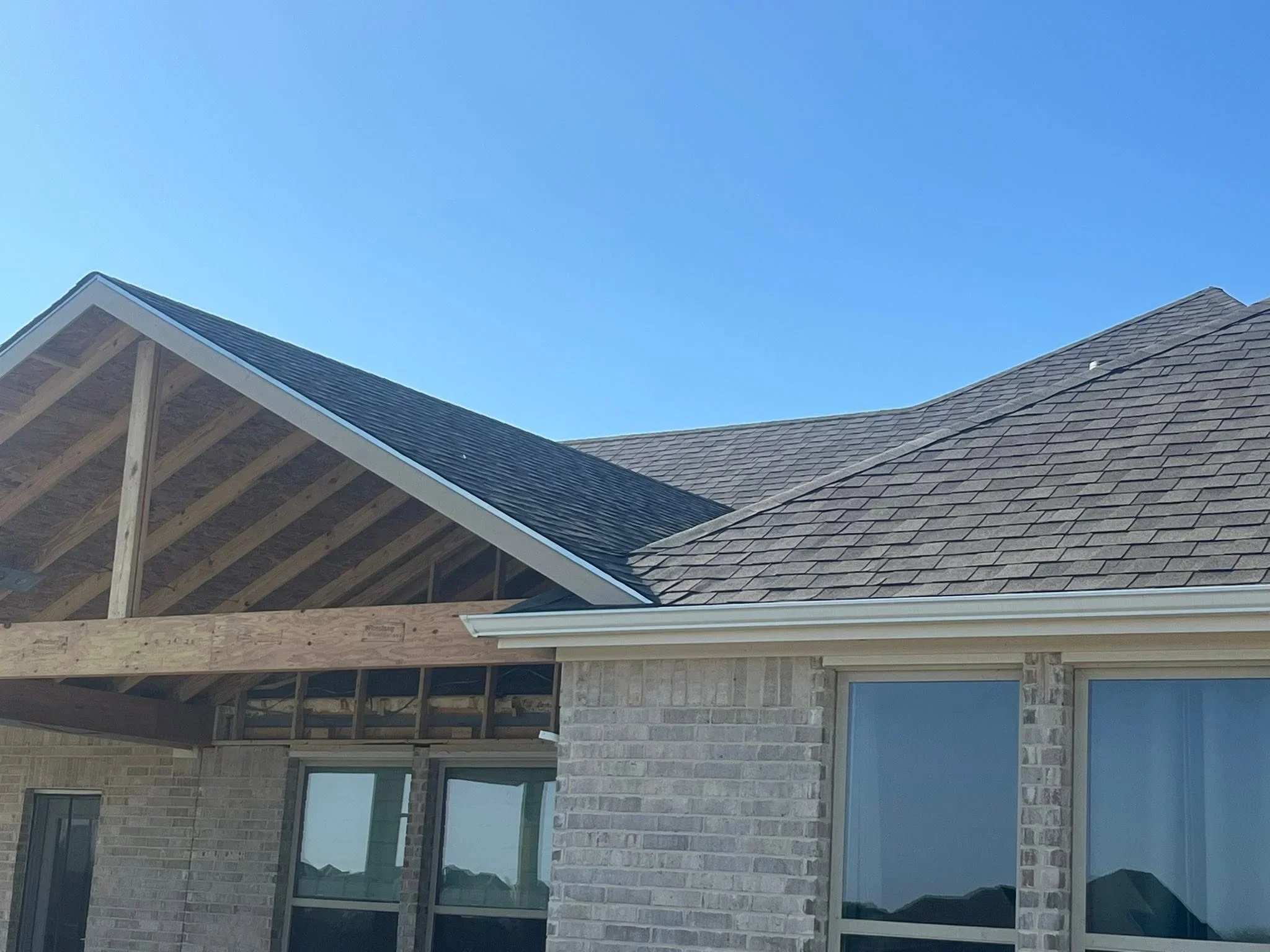 Charcoal architectural shingles, exposed wood beams, white trim, brick exterior, and large windows on residential roof