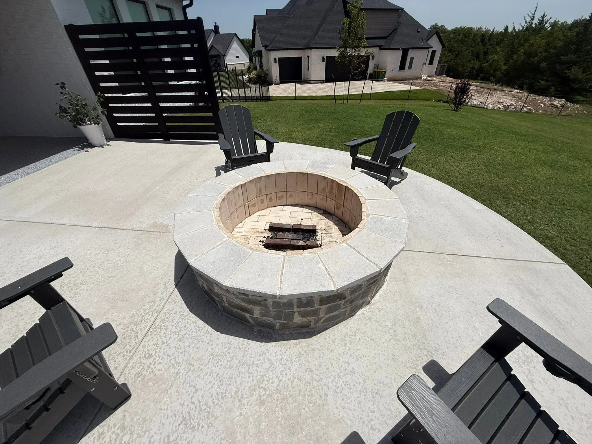 Circular stone fire pit with light cap on concrete patio, black Adirondack chairs, metal privacy screen